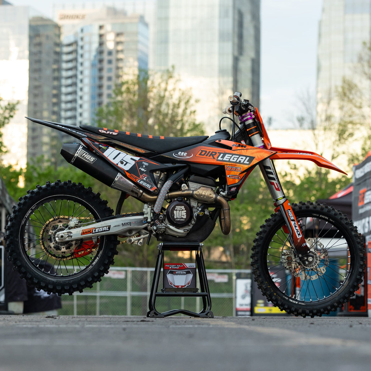 Orange and black dirt bike on a Risk Racing F.F.S. stand with cityscape in the background
